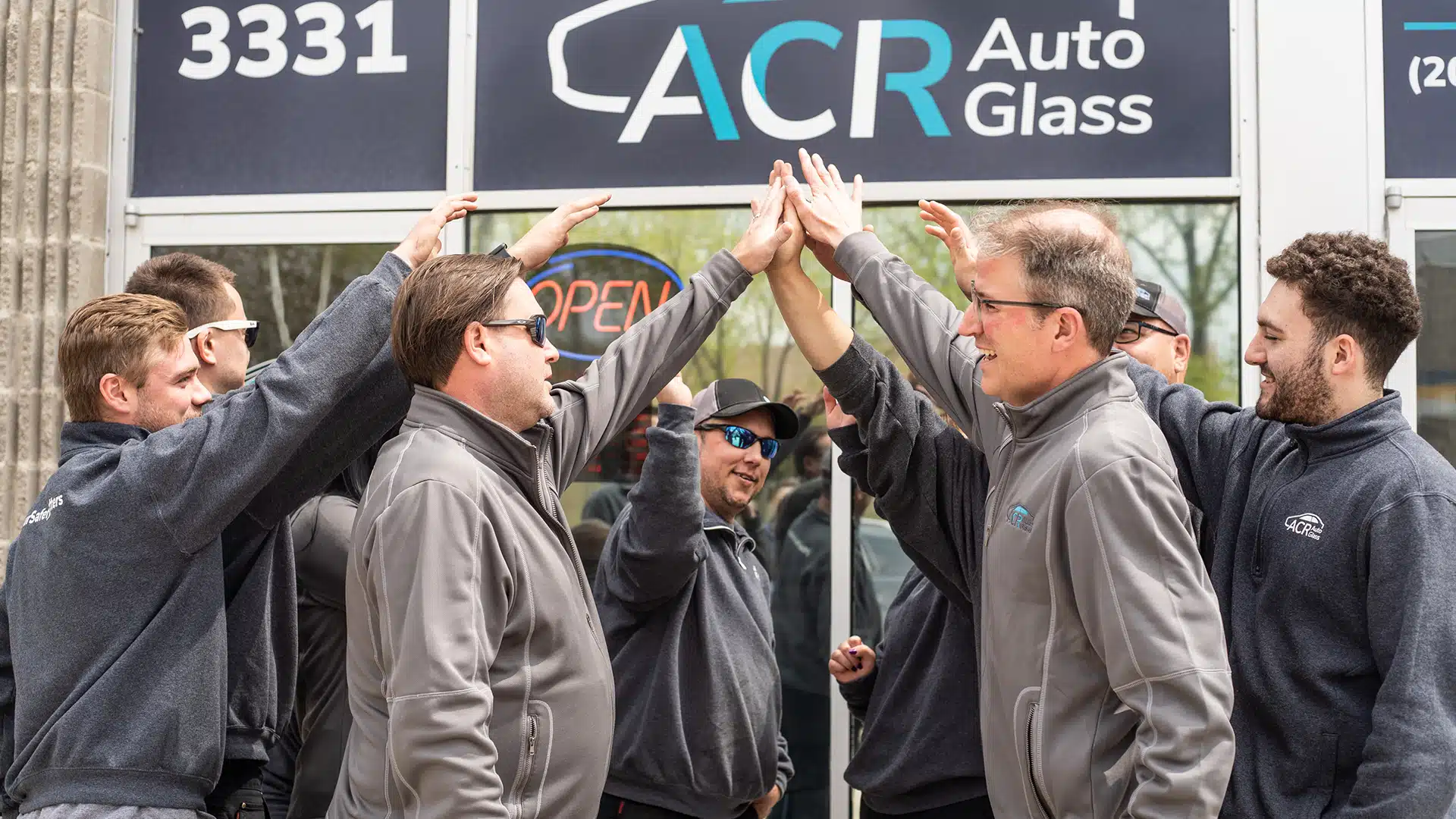 A group of people in matching gray jackets smiling and high-fiving in front of a storefront with a sign that reads ACR Auto Glass. The atmosphere appears positive and team-oriented.