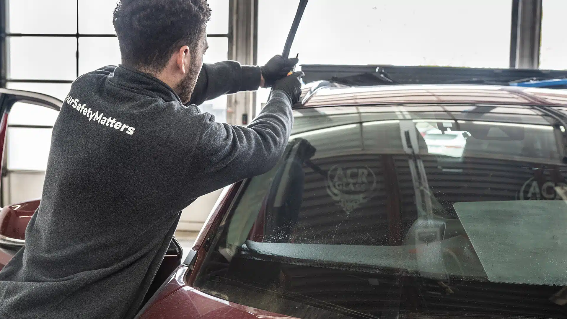 A person wearing gloves and a sweatshirt labeled “#SaferMatters” works on replacing or repairing the windshield of a maroon car inside a garage.